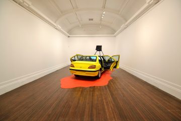 Installation by artist Heather Phillipson of yellow car on orange wooden puddle. The car has a projector built into the front bonnet that allows viewers while inside the vehicle to watch film created by artist,. Included in the exhibition Yes, Surprising Is Existence In The Post-Vegetal Cosmorama.