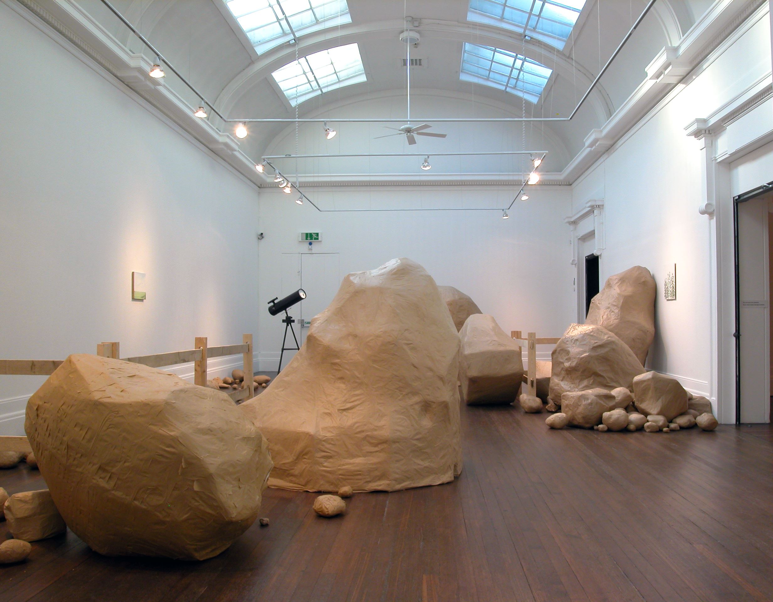 Large number of different sized paper rocks by artist Peter Liversidge. The rocks are made in a uniformly brown parcel paper colour and range from small fist sized rocks to large boulders. Included in the exhibition Peter Liversidge: What You’d Expect.