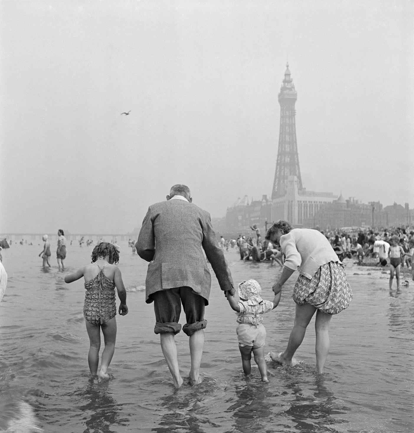 Photograph showing a family of tourists paddling in the sea with The Blackpool Tower in background. The photograph was taken by John Gay in 1949.