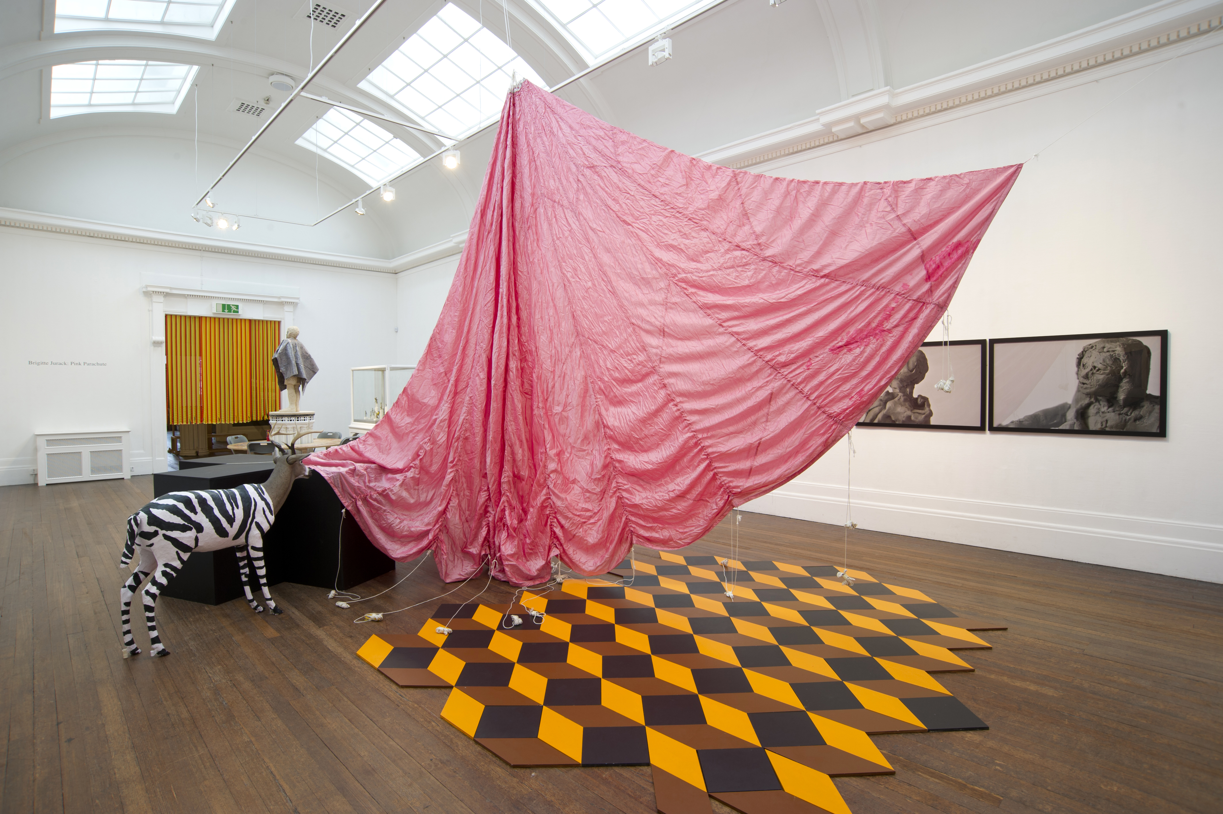 Various artworks, objects include large pink parachute, tessellated flooring, a stuffed goat/zebra hybrid, tasseled door curtain and photographs of clay faces. Included in the exhibition Brigitte Jurack: Pink Parachute.