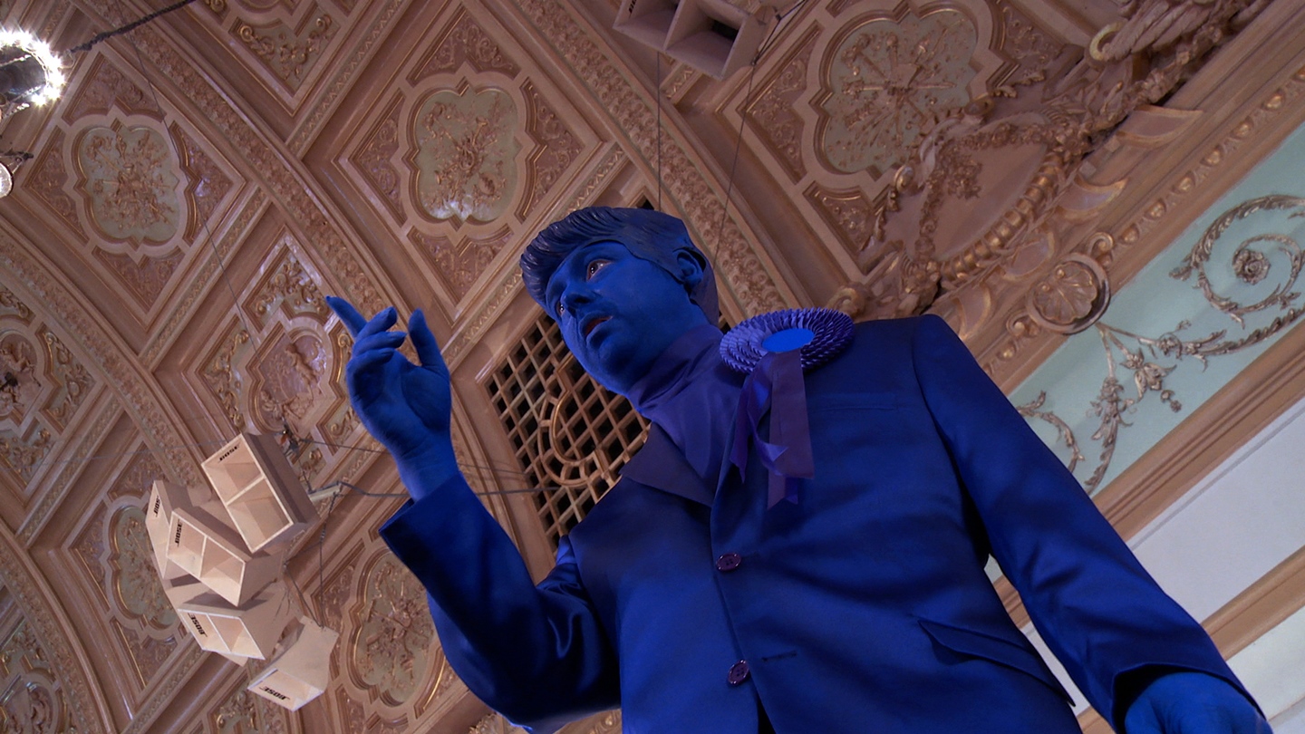 Image from film The Unspeakable Freedom Machine by artist Jennet Thomas. The image is of a male figure dressed completely in blue, wearing blue face make up. the figure is shot from below against the backdrop of The Blackpool Winter Gardens Empress Ballroom.