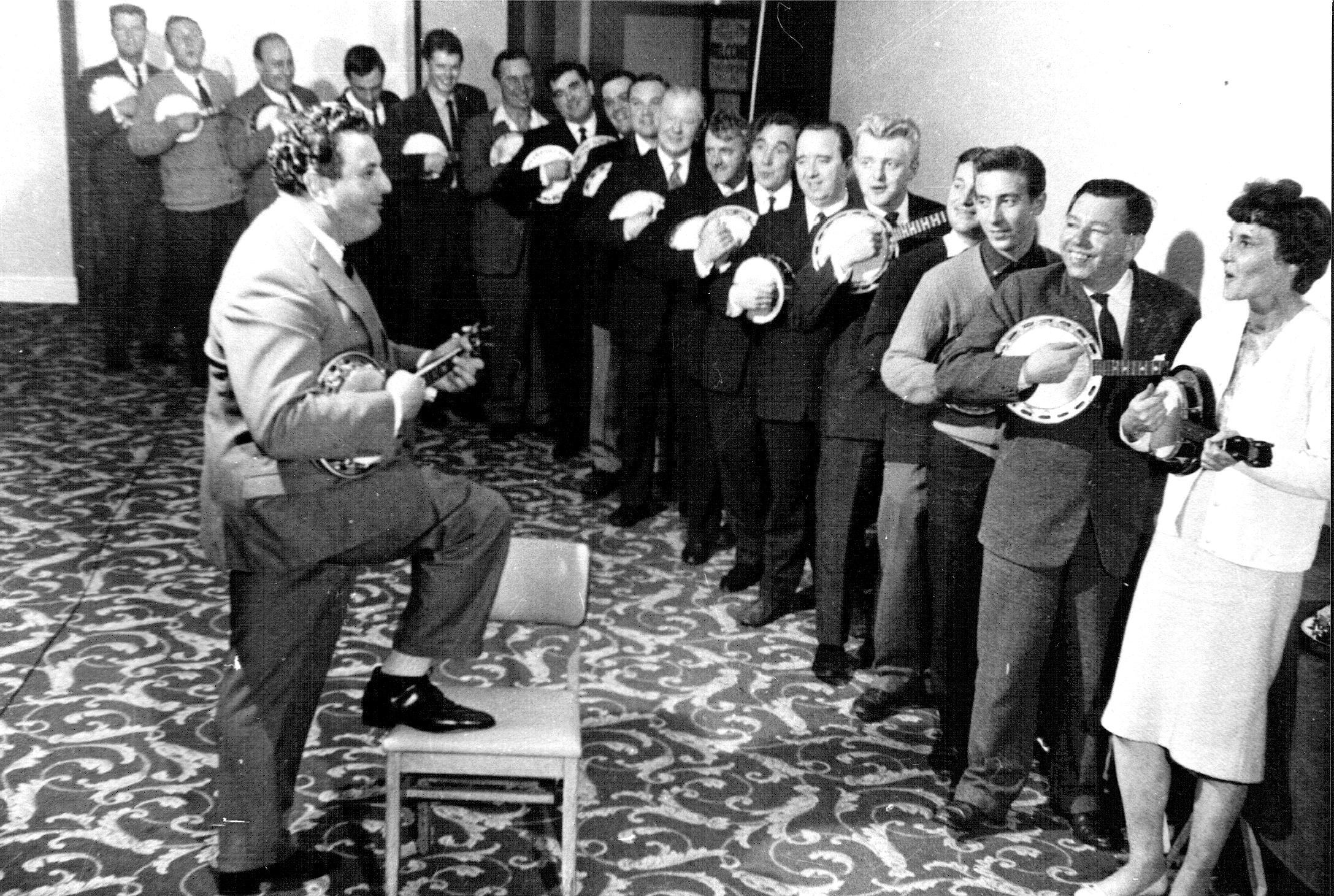 A line of ukulele players including George Formby facing a ukulele player with his foot on chair. Included in the exhibition Fan Club And My Generation.