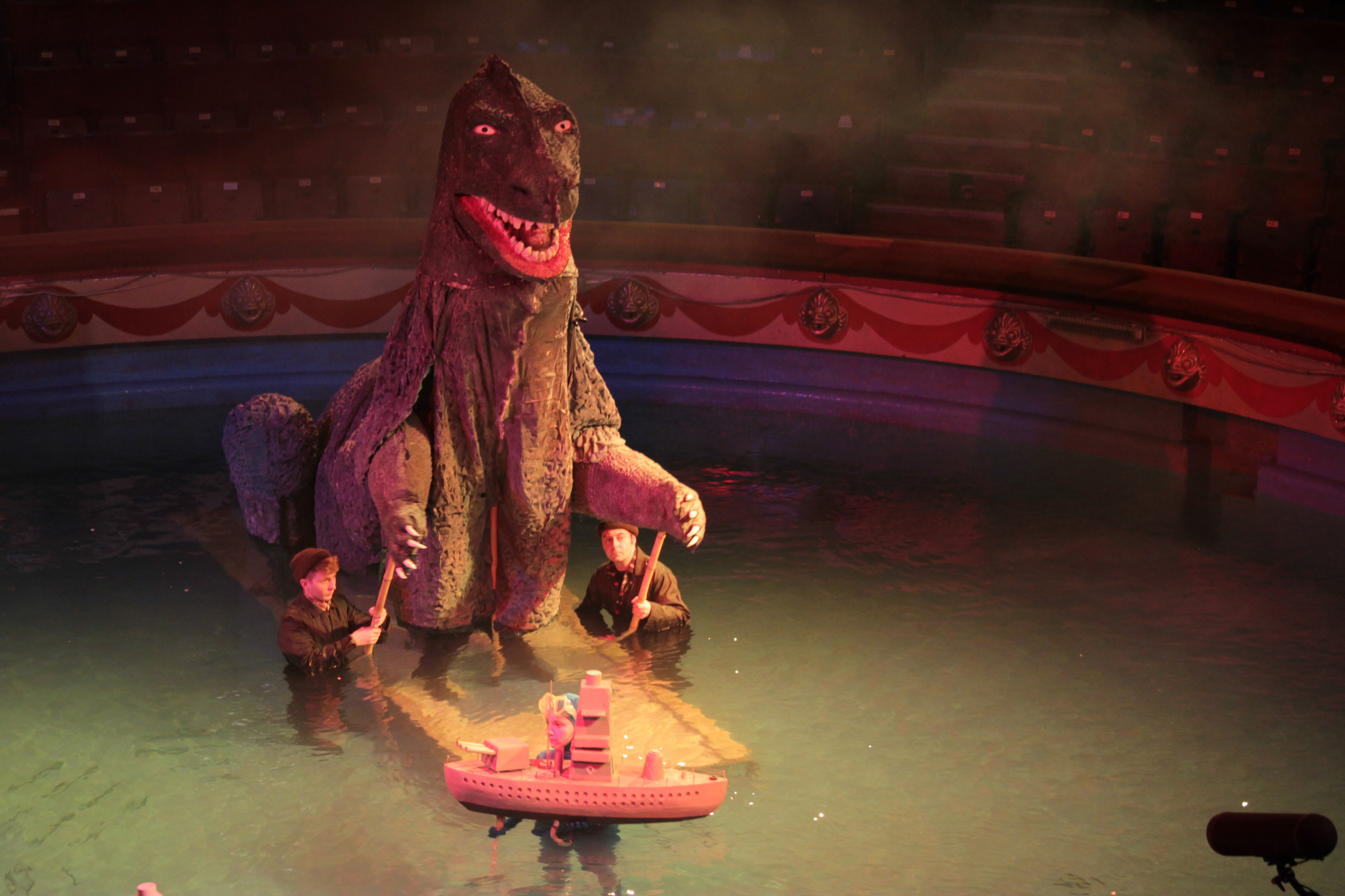 Giant sea monster puppet in the water filled Blackpool Tower Circus ring. The monster was featured in a film created by artist Serena Korda. The monster's arms are operated by 2 male figures in overalls while a female figure in wetsuit operates a floating model of a battleship. The monster and film were included in the exhibition Aping The Beast.