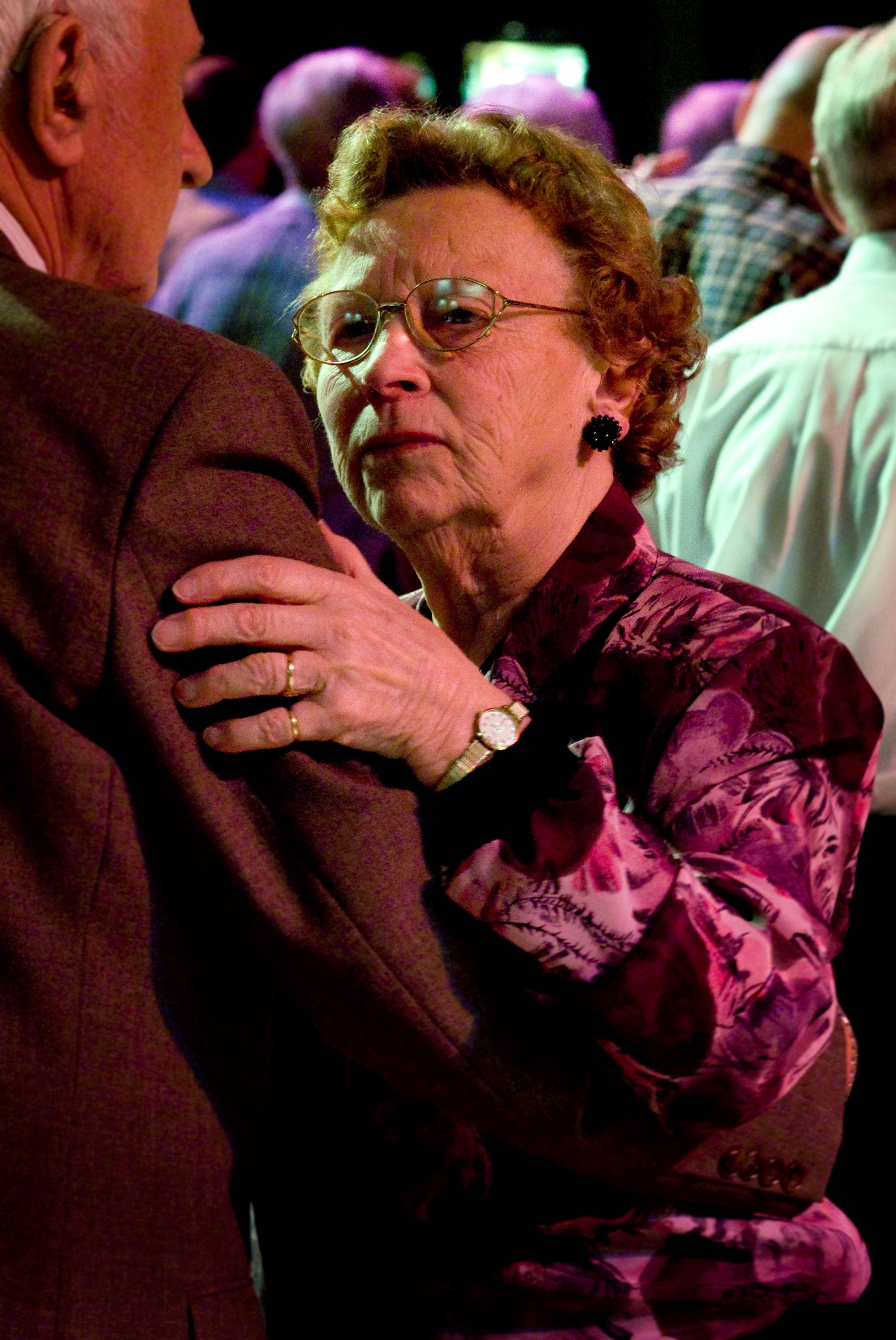 Elderly couple dancing in The Blackpool Tower Ballroom. The male dancer is wearing a brown suit and is facing away from camera, the female dancer is wearing a dark red rose pattern, and is looking into the camera. The image was taken by photographer Linzi Cason and included in the exhibition Civic Photography.