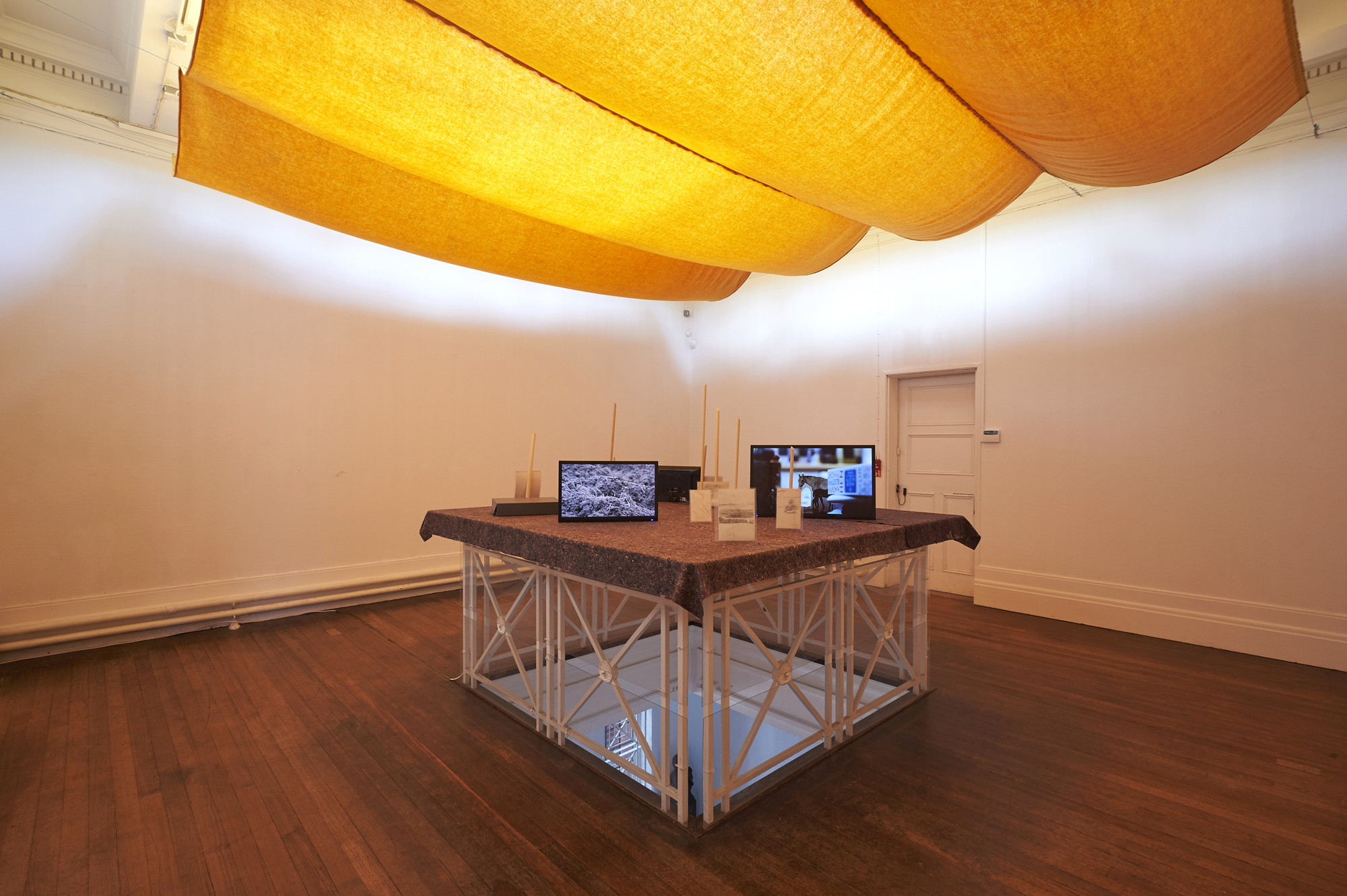 The Grundy Art Gallery’s upstairs rotunda space featuring installation of various lengths of sticks and three screens showing videos on blanketed display board created over The Grundy Art Gallery’s railing bordered dome viewing floor area.The dome has been blocked from view by hanging curtains. Included in the exhibition Fig-Futures.