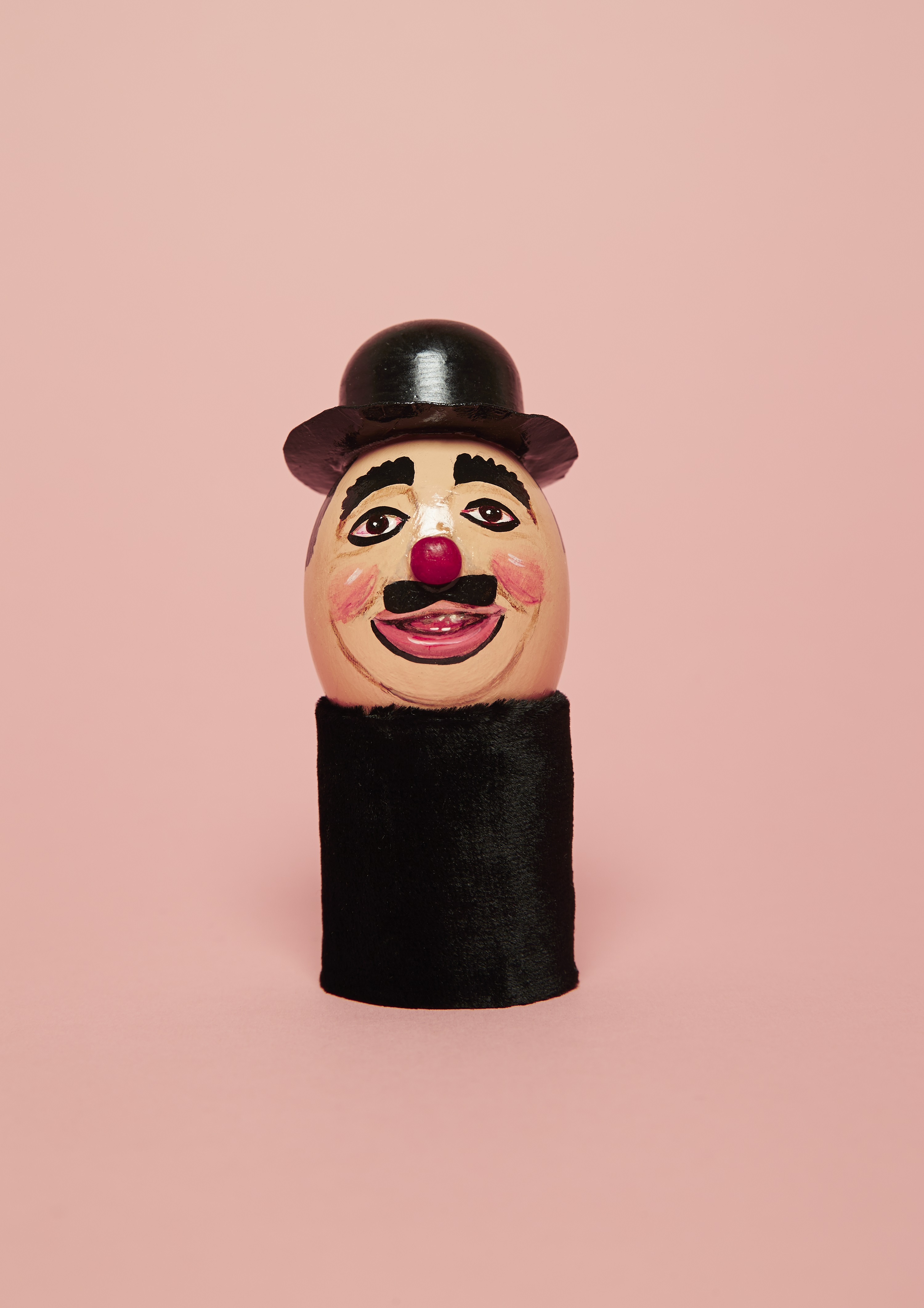 Egg painted with clowns face. The egg has the make up of clown Charlie Cairoli painted on it, it wears black bowler hat and is sitting on black tube in front of peach background. The Photograph is part of the clown egg register, a visual history of registered clown performers make up by Luke Stephenson Photograph included in the exhibition A Clown Walks Into A Gallery…