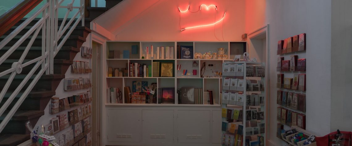 Shop area of The Grundy Art Gallery featuring a selection of cards and other items. above rear of shop area is a red neon artwork of letters and line, the letters say UM. The artwork is by artist Noel Clueit, and it responds to a similar neon artwork on permanent display over The Grundy Art Galleries entrance doorway.