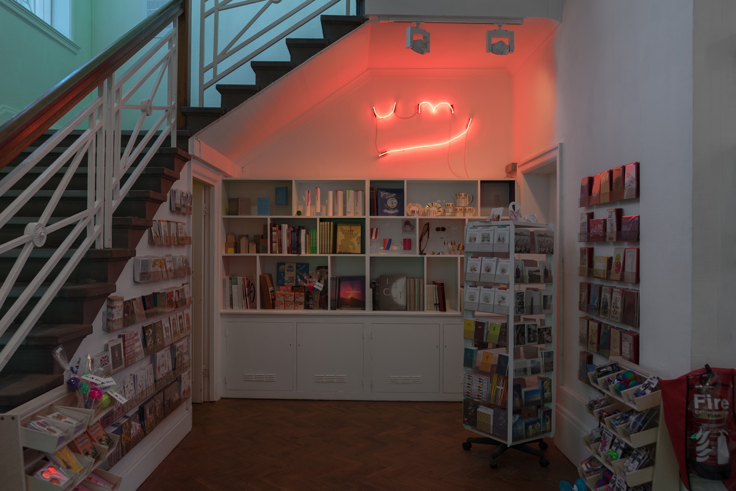 Shop area of The Grundy Art Gallery featuring a selection of cards and other items. above rear of shop area is a red neon artwork of letters and line, the letters say UM. The artwork is by artist Noel Clueit, and it responds to a similar neon artwork on permanent display over The Grundy Art Galleries entrance doorway.